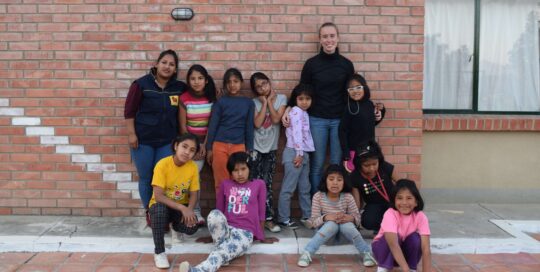 Die Freiwillige Felicitas steht an eine Wand gelehnt mit den Kindern aus dem Kinderdorf zusammen